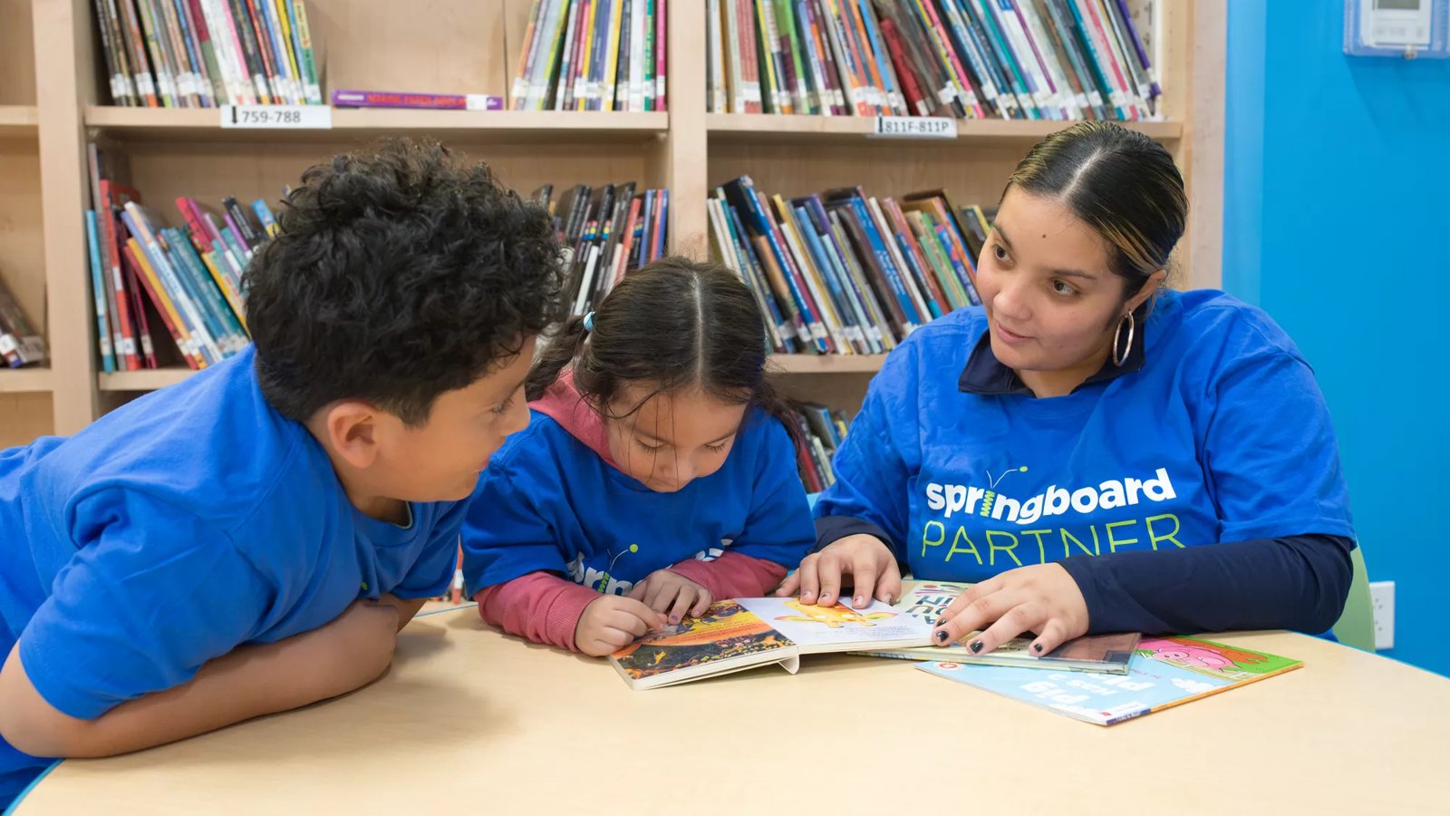 Two children and a woman read books in a library.