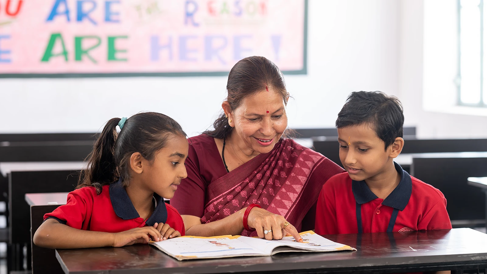 A teacher reads a book with two students in a classroom.