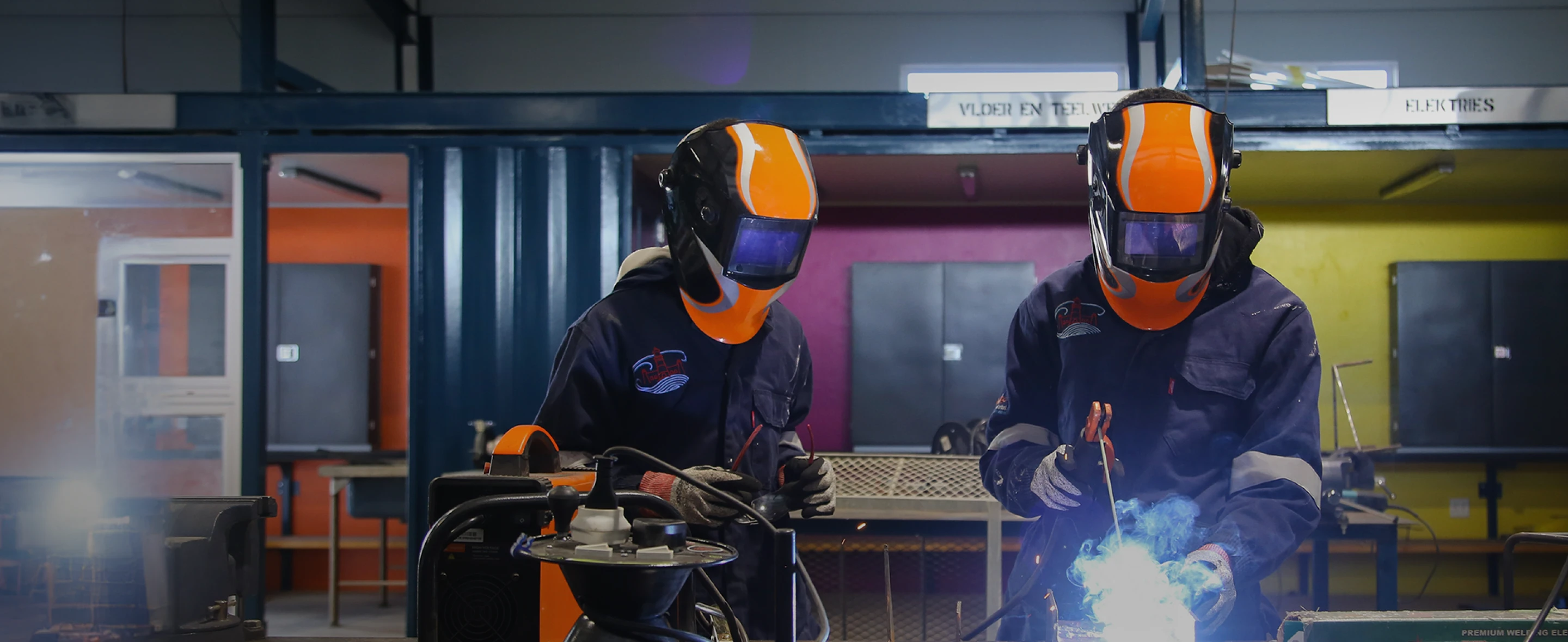 Two people wearing welding helmets in a factory setting.