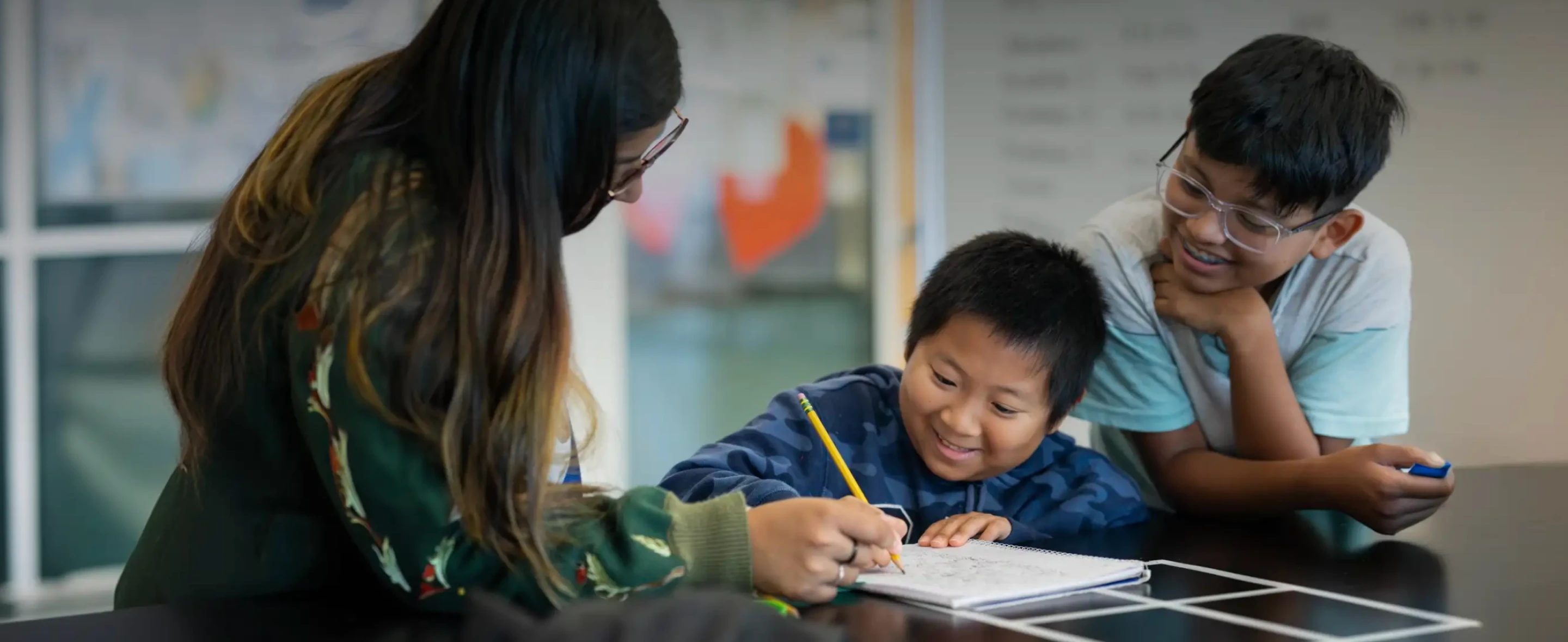 One adult and 2 children are gathered around a table; one child is writing in a notebook, with the others observing and smiling.