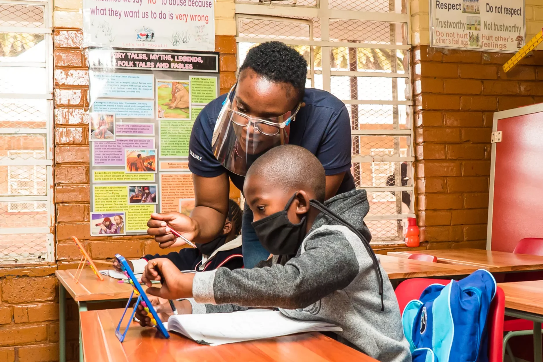 A teacher in a mask works with a child in a mask in a South African classroom.