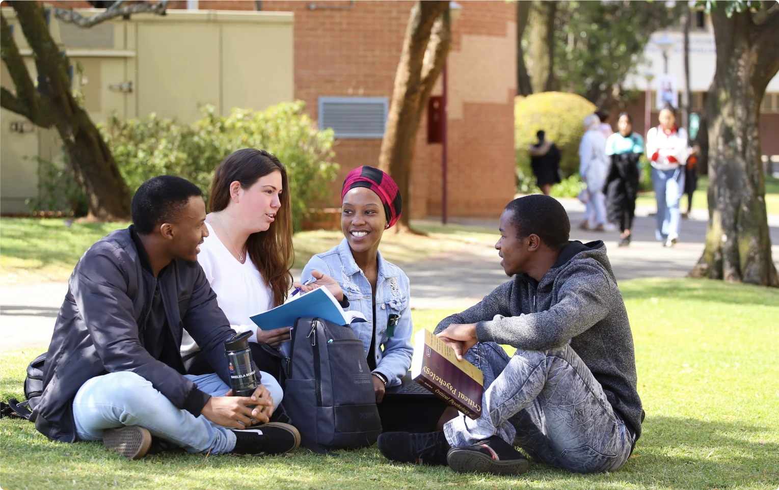 Four students sit on the grass, chatting on a university campus.