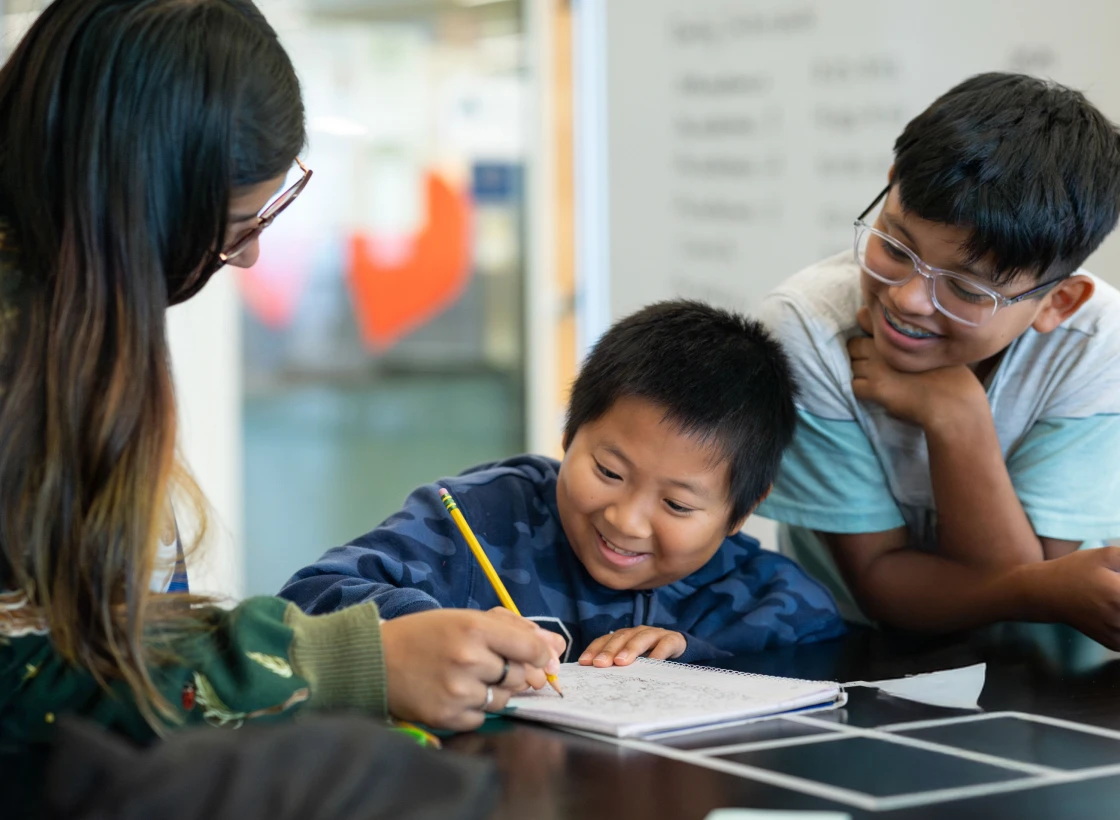 One adult and 2 children are gathered around a table; one child is writing in a notebook, with the others observing and smiling.