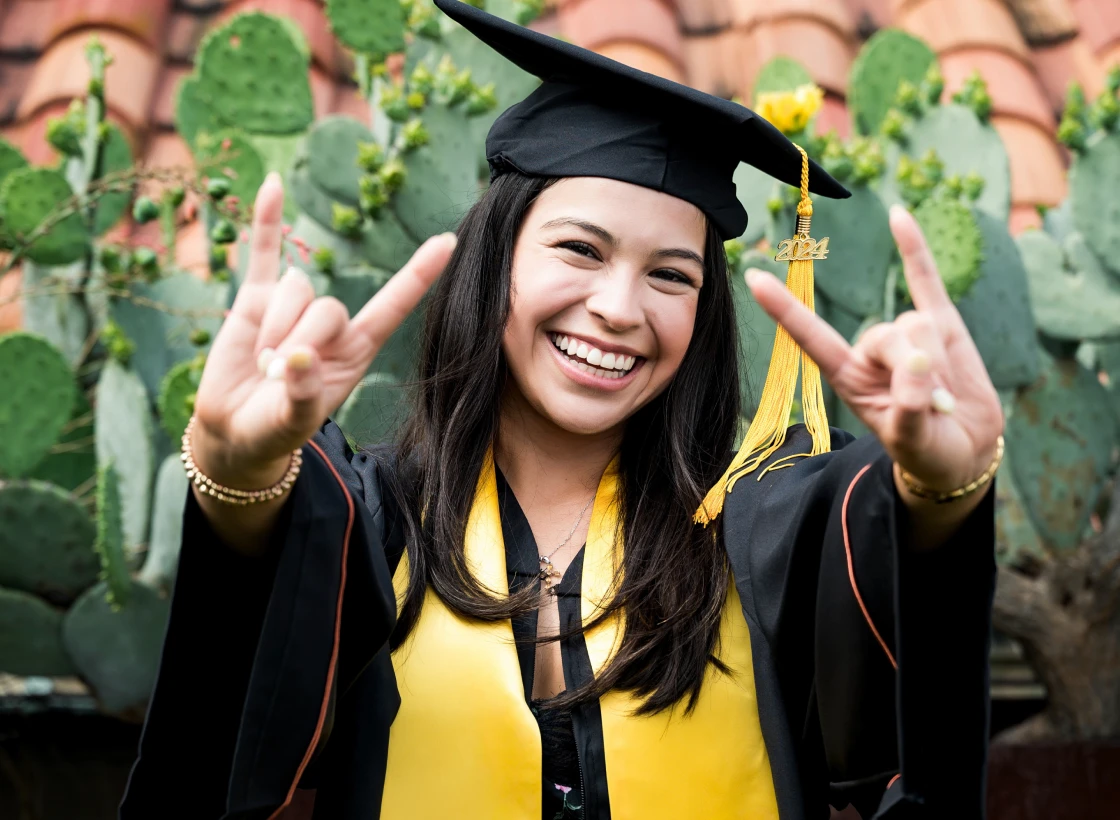 Woman in graduation gown smiling and making hook 'em horns sign.