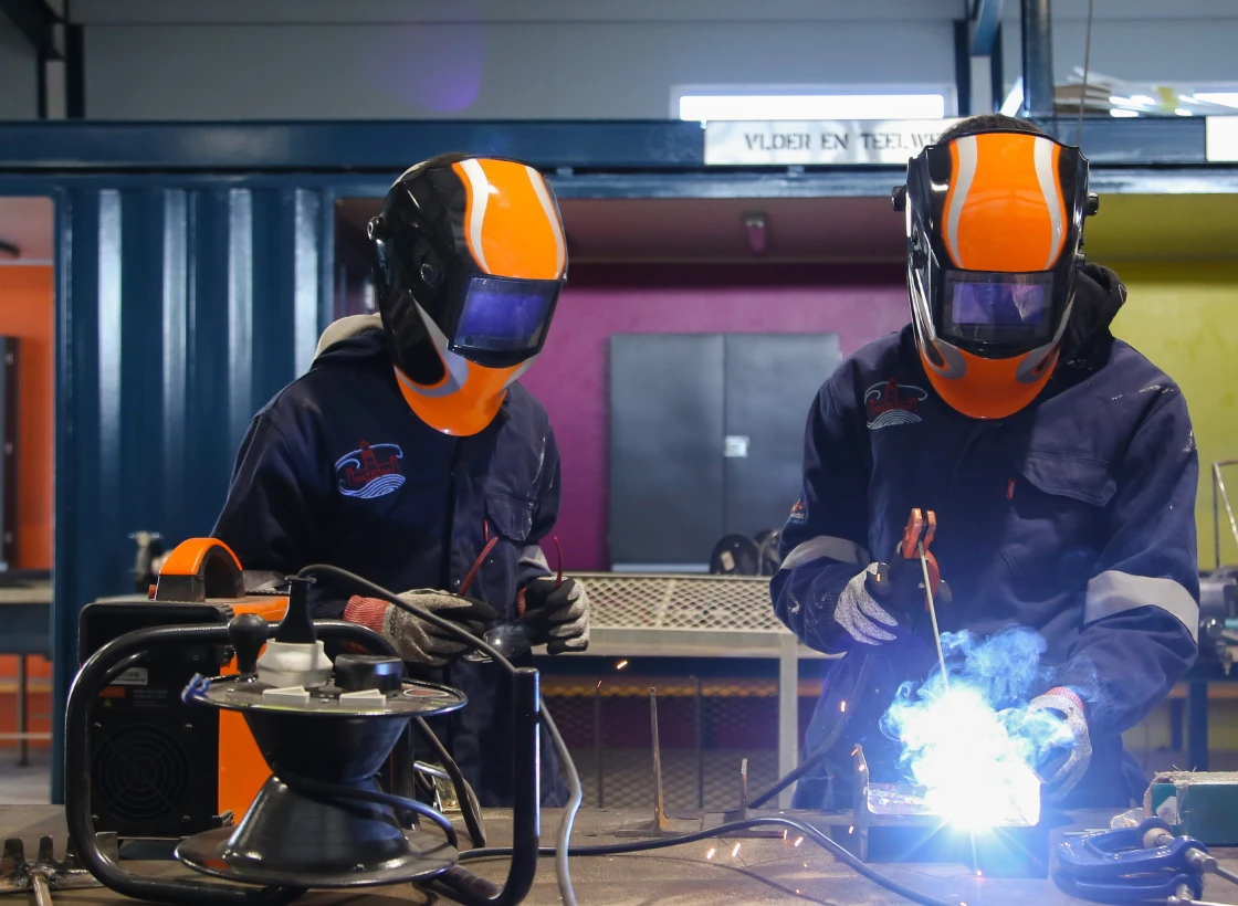 Two people wearing welding helmets in a factory setting.