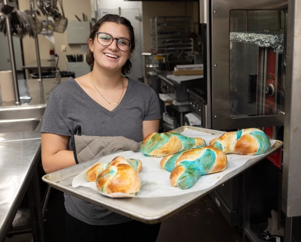 A woman holds a pan of freshly baked challah while standing in a kitchen.