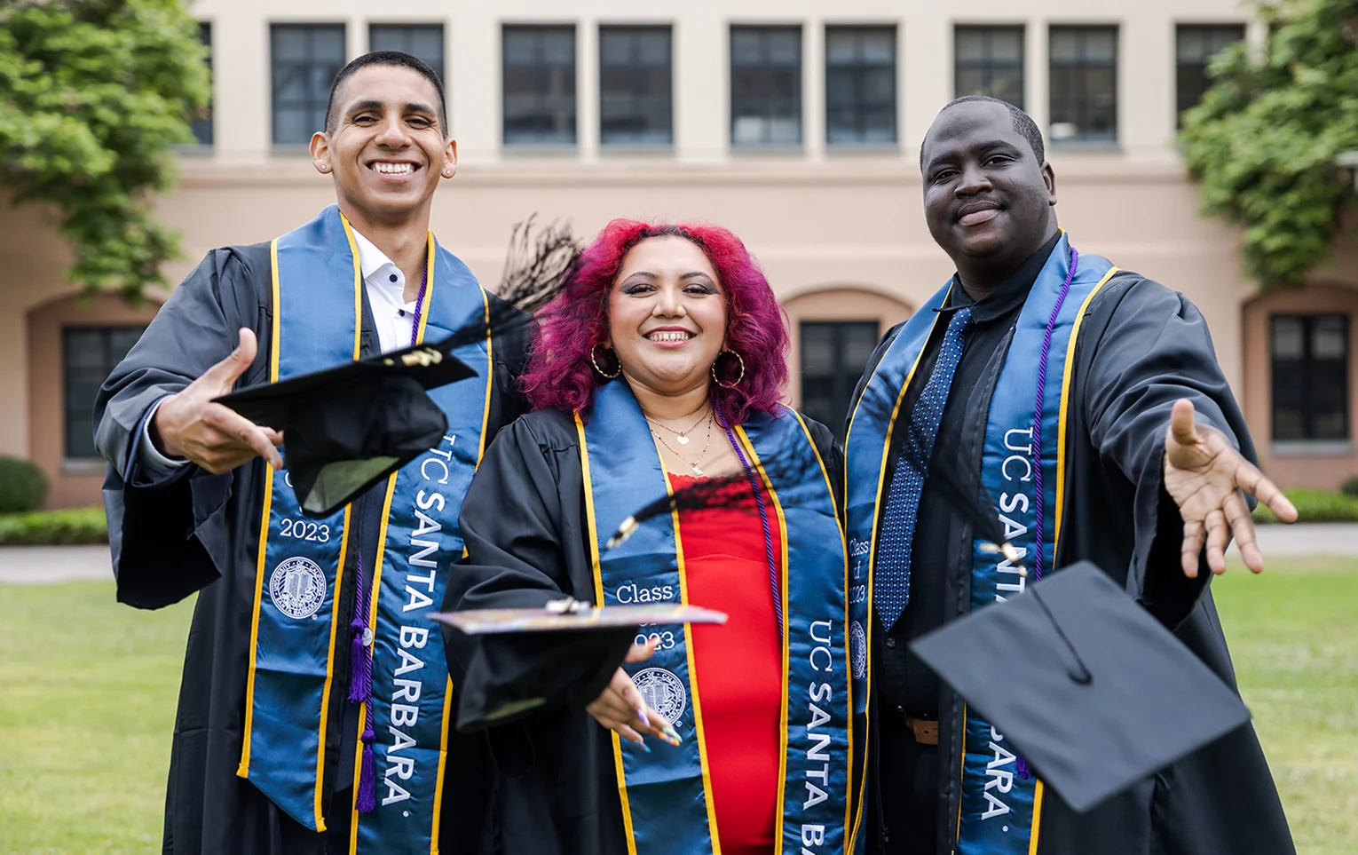 Three university graduates smile while throwing their graduation caps toward the camera.