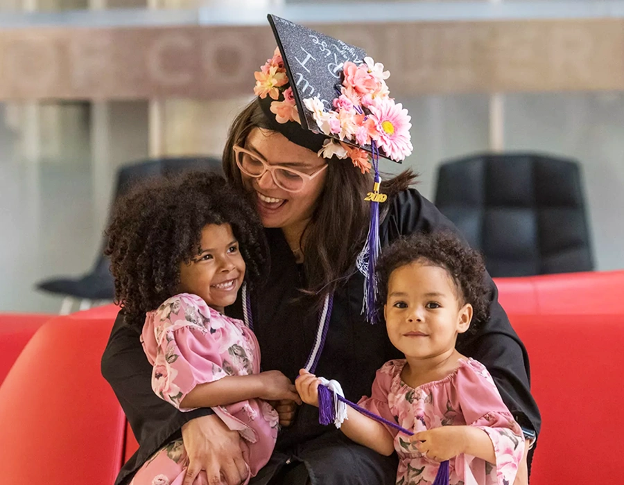 A woman in a graduation cap and gown hugs two small children.