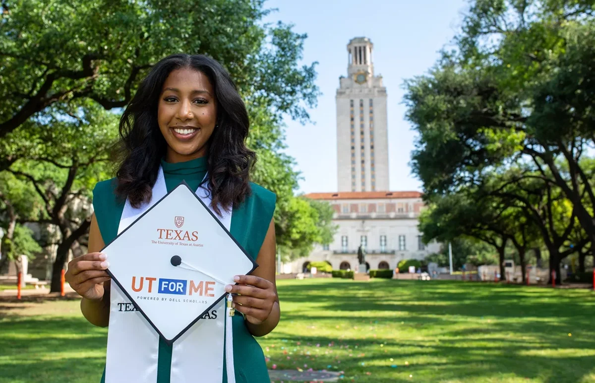 A Dell Scholar graduate stands in front of the UT Tower with her UT for Me graduation cap in hand.